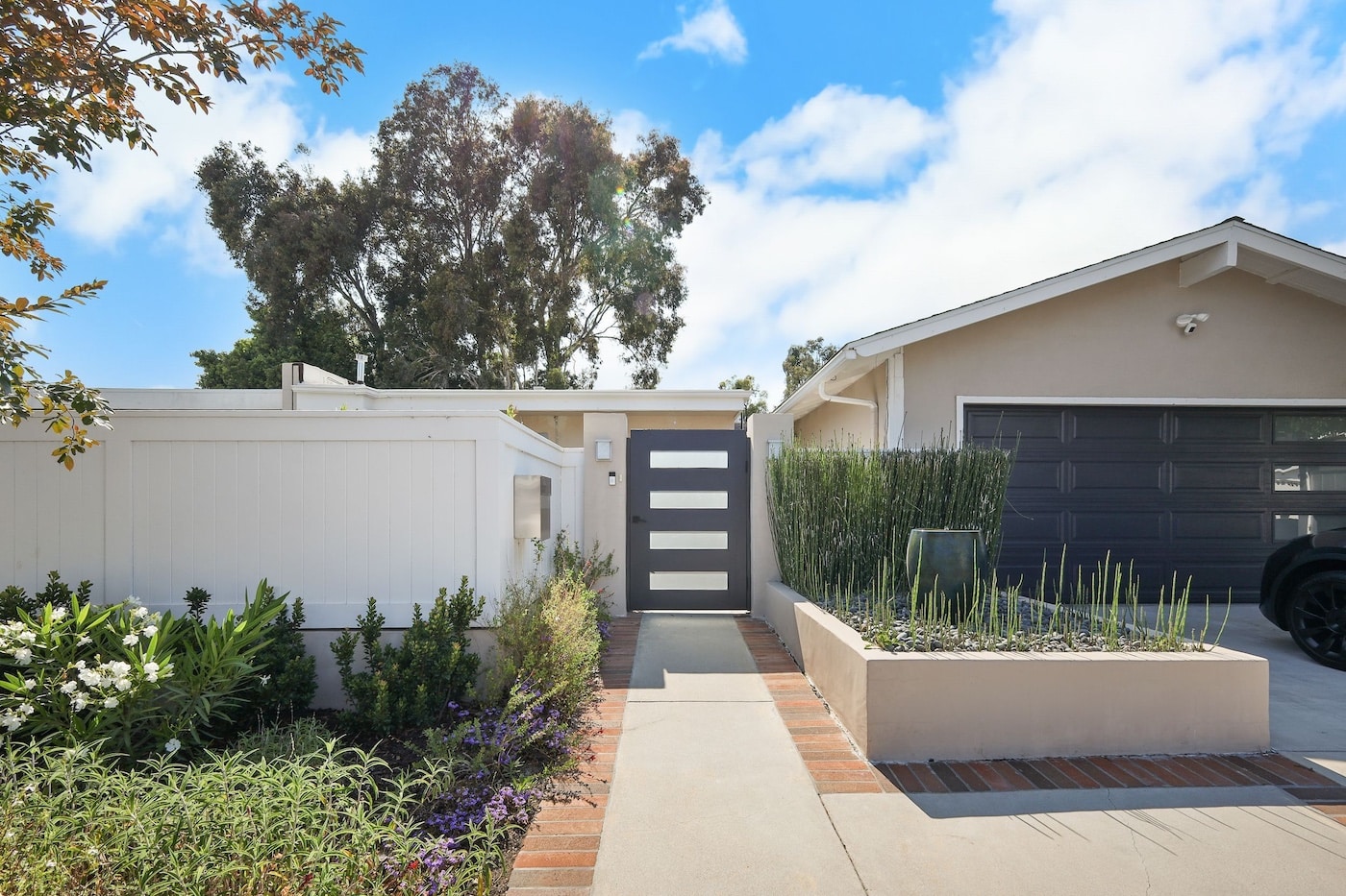 Modern house with a dark garage door, white fence, and lush garden under a clear blue sky.
