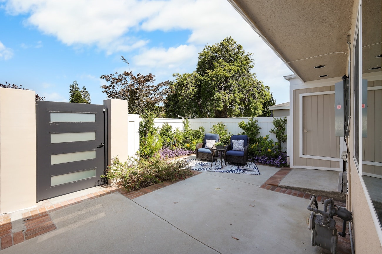 Cozy patio with two blue chairs, small table, white fence, and lush greenery under a blue sky.