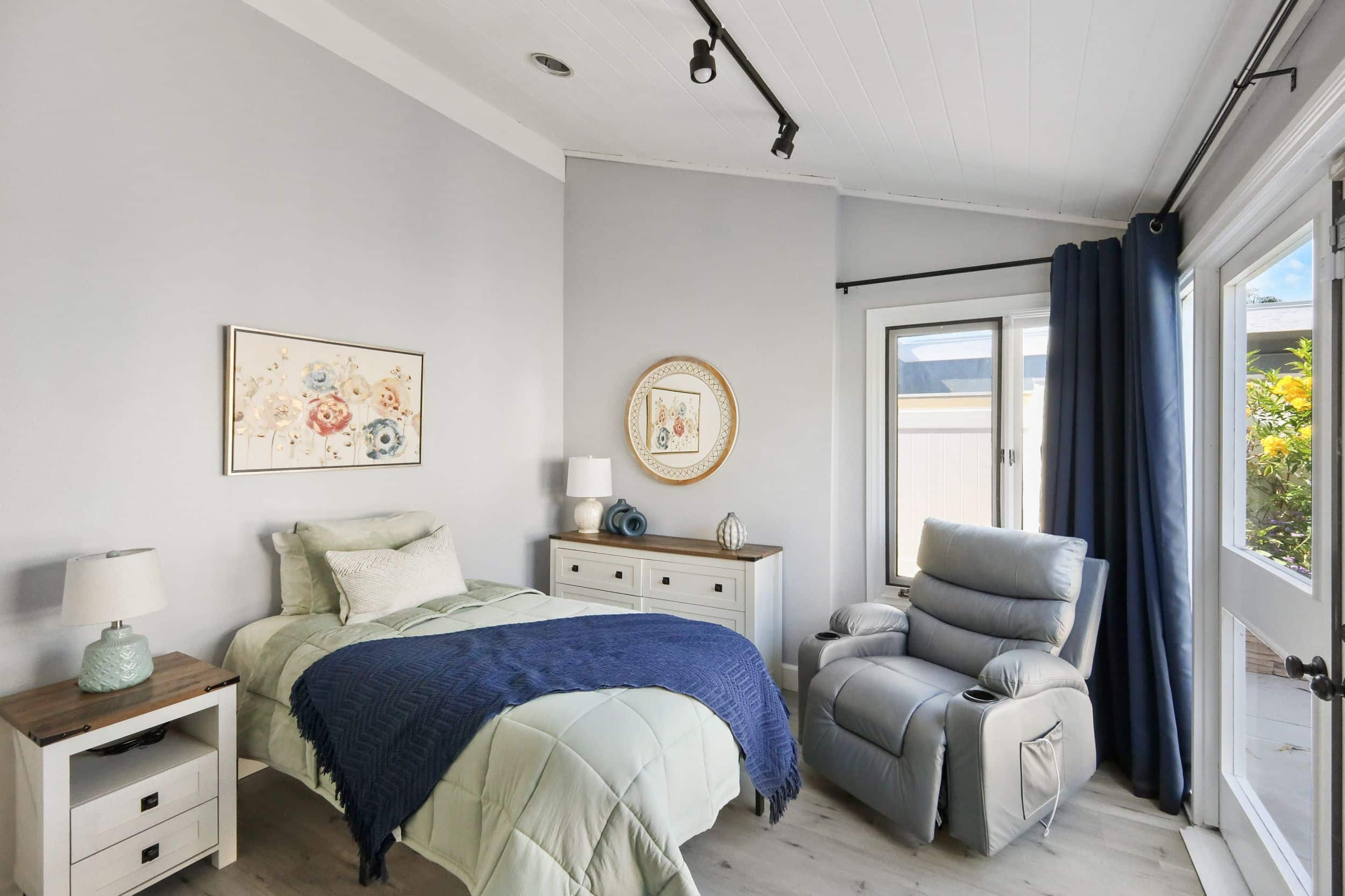 Cozy bedroom with bed, recliner, blue blanket, and floral artwork on walls.
