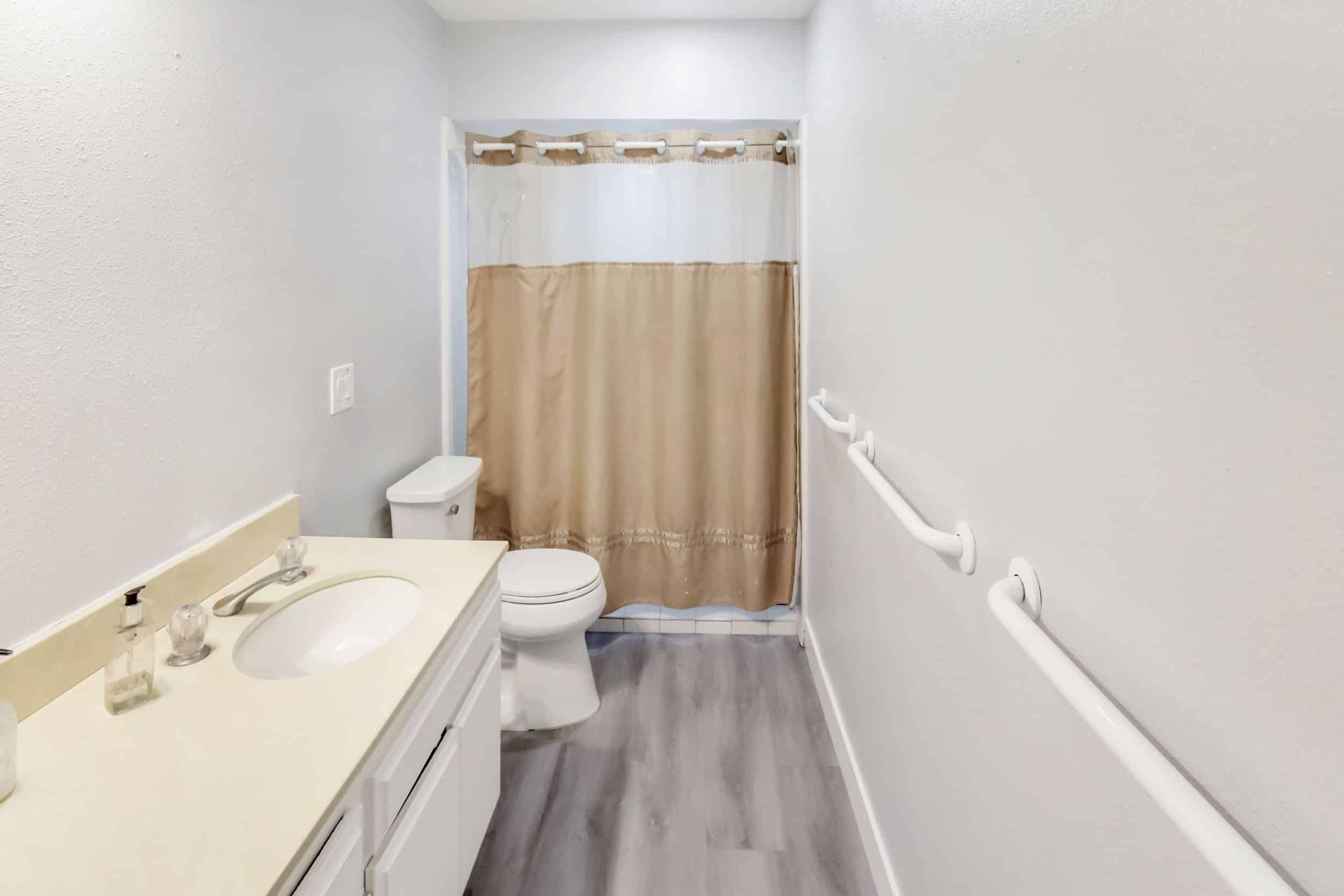 Accessible bathroom with beige shower curtain, grab bars, and light gray flooring.