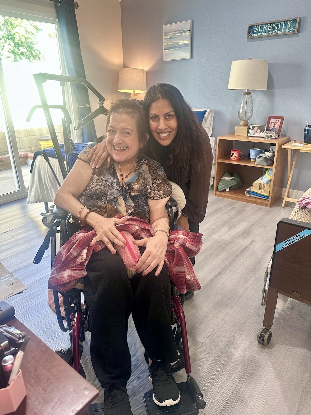 A smiling woman in a wheelchair with a blanket and a person standing beside her in a cozy room.