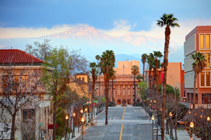 City street view with palm trees, buildings, and mountains in the background at sunset or sunrise.