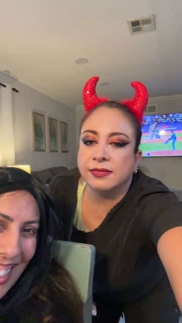 A woman wearing red devil horns poses indoors with a baseball game on TV in the background.