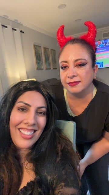 Two women in costumes; one with red horns and makeup, smiling indoors.